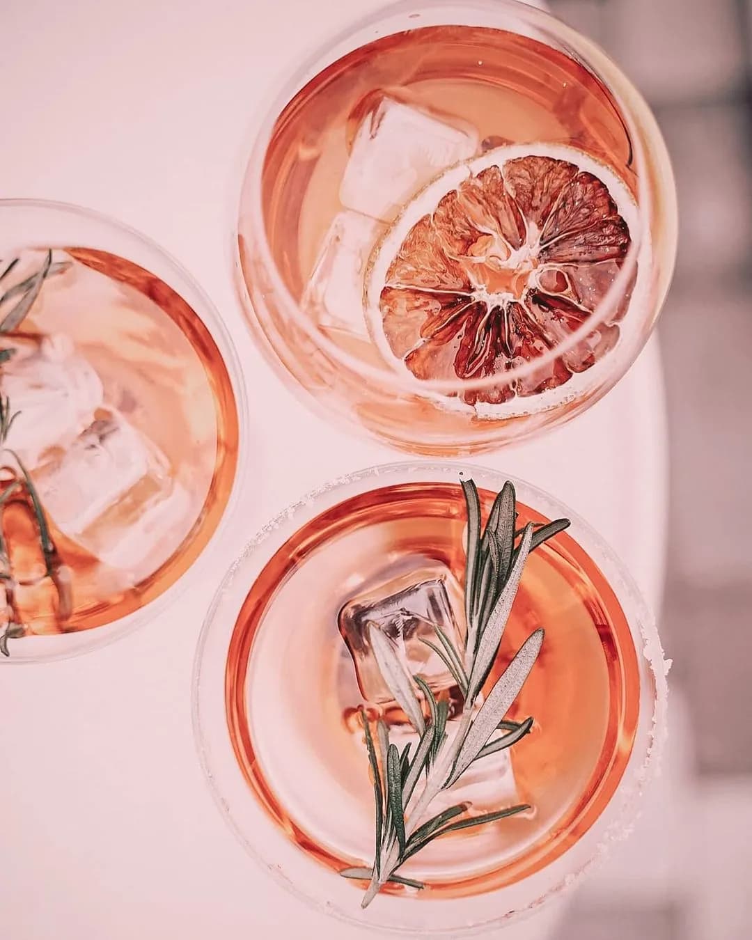 Top-down view of three glasses filled with a light pink beverage, each with ice cubes. One drink is garnished with a slice of dried citrus fruit, while the other two are adorned with sprigs of rosemary. The drinks are placed on a white surface.