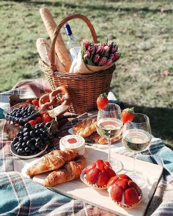 A picnic scene with a woven basket containing baguettes, a bottle of white wine, and a bouquet of tulips. On the blanket and board, there are croissants, grapes, strawberries, soft cheese, strawberry tarts, pretzels, and two glasses of white wine.