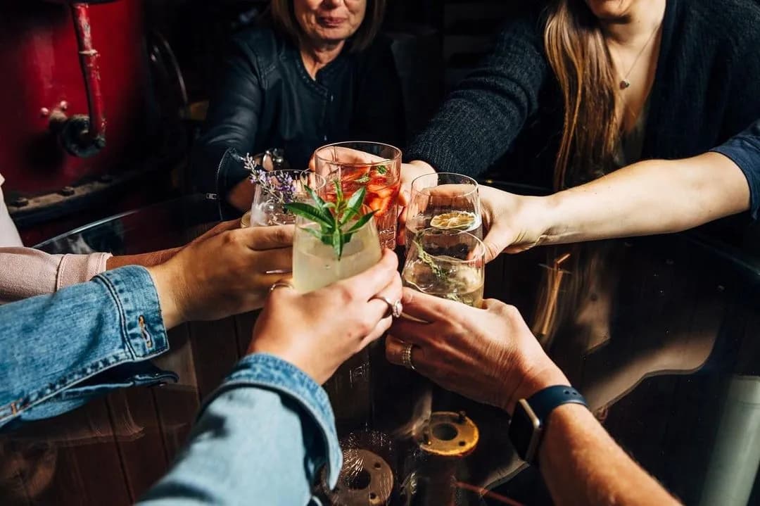 Five people clink glasses together in a toast around a dark table. Each person holds a different drink, including a cocktail with a garnish, a red drink with ice, and a glass of white wine. One person is wearing a denim jacket, and another is wearing a smartwatch.