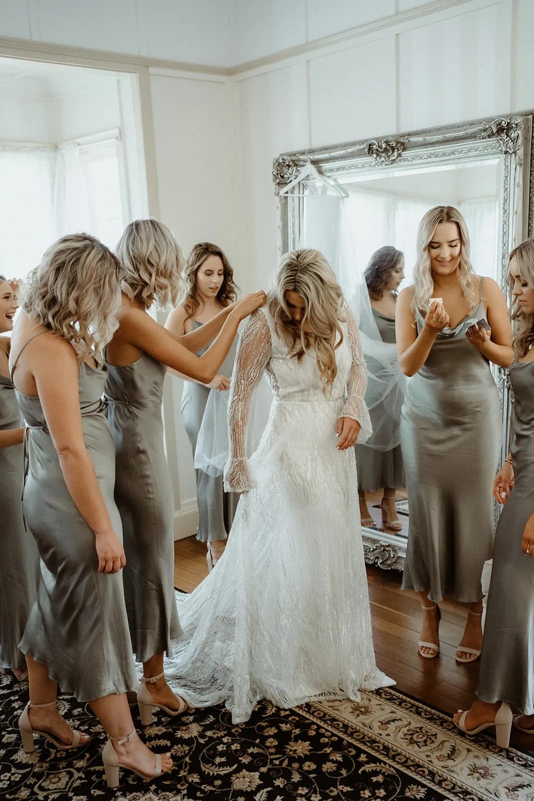 A bride in a white gown is being assisted by bridesmaids wearing matching silver dresses. They are gathered in a well-lit room with a large ornate mirror and wooden floors, preparing for the wedding ceremony. The atmosphere is joyful and intimate.