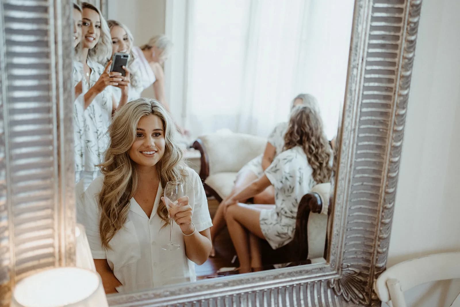 A woman with long, blonde hair, dressed in white, smiles and holds a glass of wine while looking into a mirror. In the background, three other women in matching white attire, one holding a phone, are also reflected in the mirror. They're in a cozy, well-lit room.