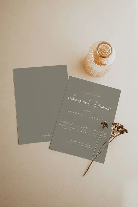 Two gray cards for a rehearsal dinner lay on a beige surface beside a small brown glass vase and dried flowers. One of the cards has text detailing the event, including names, date, and location, while the other card is blank.