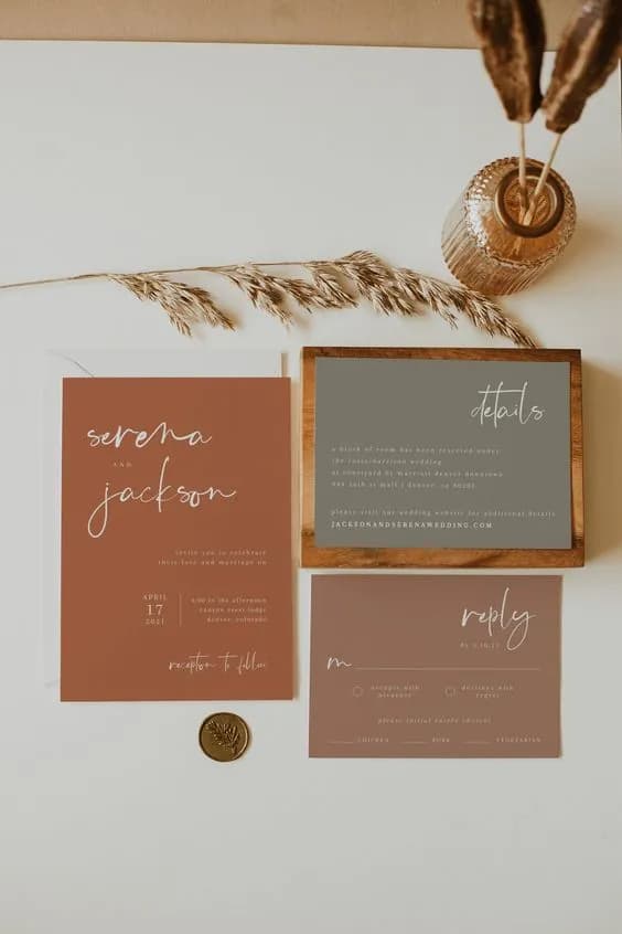 A flat lay of wedding stationery items on a light surface, including an invitation card, details card, and RSVP card. The invitation reads "Serena and Jackson," and the background colors are earthy tones. Decorative dried grasses and a wax seal are also present.