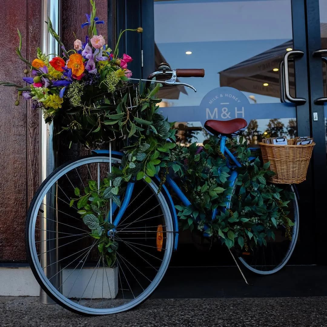 A blue bicycle adorned with green foliage and colorful flowers is parked in front of a café entrance. The front basket contains a vibrant floral arrangement, and the rear basket is empty. The café's door features the logo "M&H" on the glass.