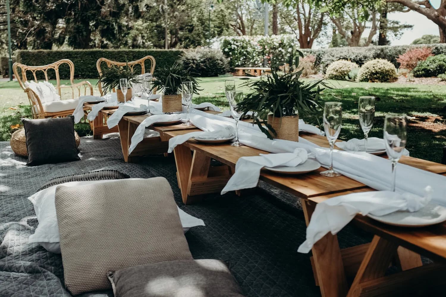 A picnic setting with two wooden benches and cushioned seats around a low wooden table adorned with white cloths and greenery. Champagne flutes and plates are neatly arranged on the table. The scene is outdoors on a sunny day with trees and bushes in the background.