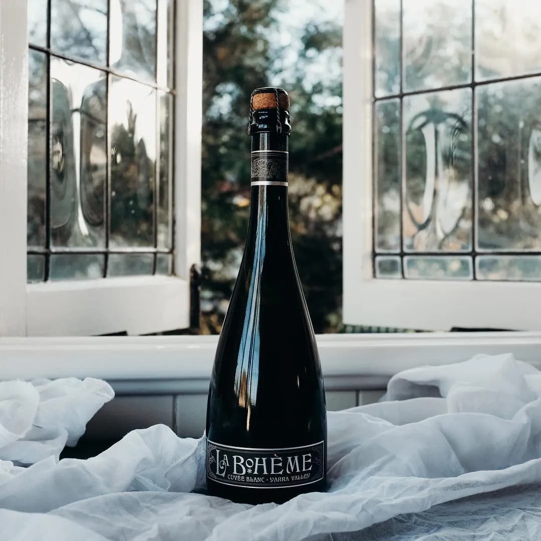 A dark bottle of La Boheme Cuvee Blanc sparkling wine stands on a white cloth, set in front of an open window with stained glass panels. Sunlight filters through the window, creating a serene and elegant atmosphere.