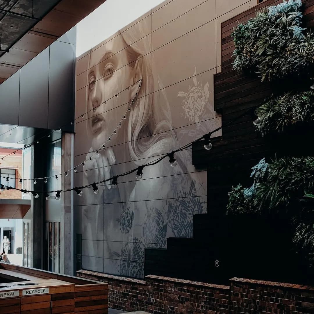 A large grayscale mural of a woman's face adorns the side of a building in an urban setting. The mural features intricate details and shades. Steps with potted plants and string lights add greenery and ambiance. A few people are seen in the background.