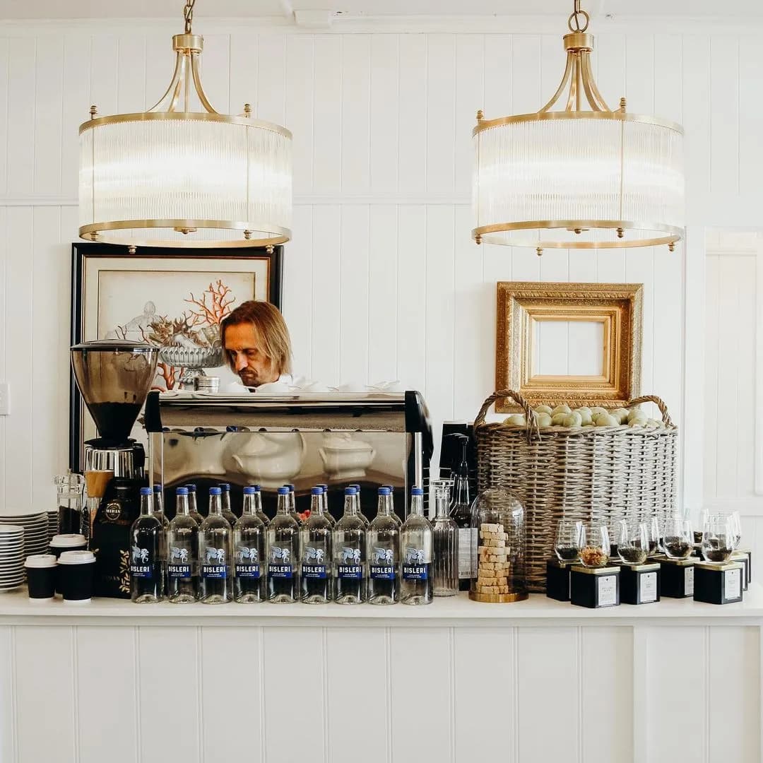 A barista prepares coffee behind a counter adorned with a coffee machine, cups, and an array of mineral water bottles. The decor features two elegant chandeliers and framed artwork on white paneled walls. A basket of green apples and jars of condiments are also present.