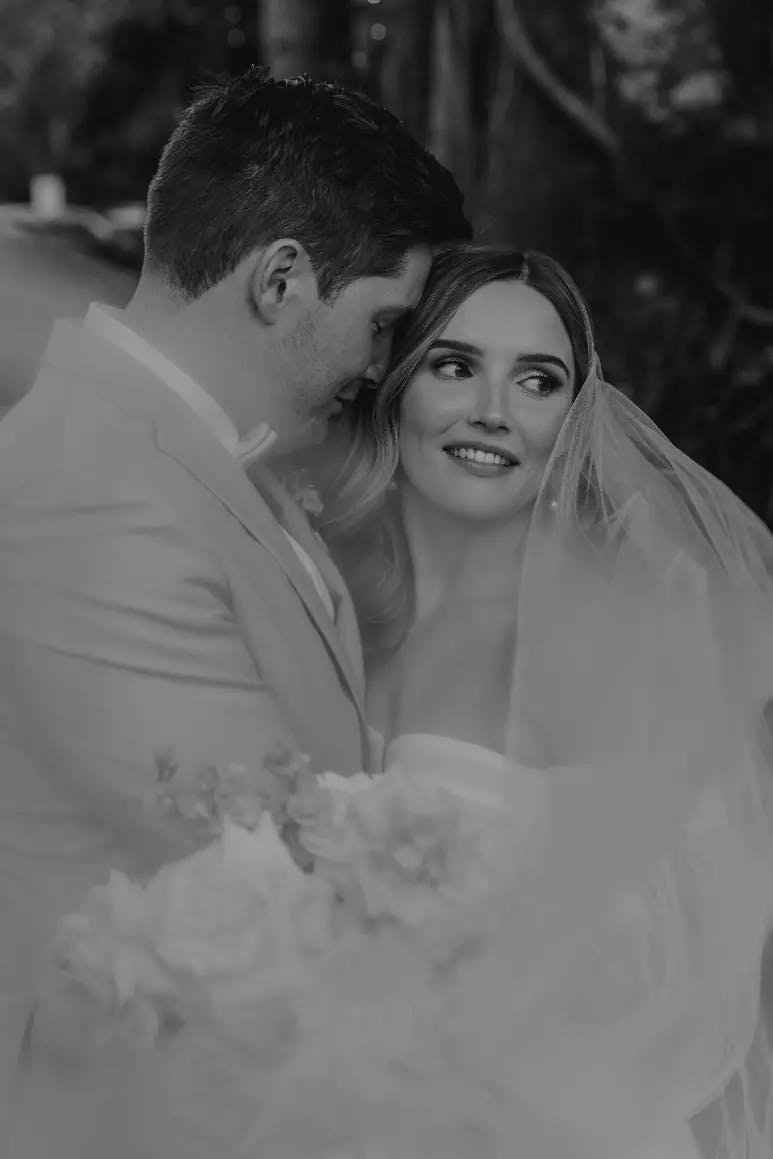 A black and white photo of a bride and groom on their wedding day. The groom, wearing a suit, lovingly embraces the bride, who is in a white wedding dress and veil. The bride looks away, smiling softly. They are surrounded by a serene, natural background.