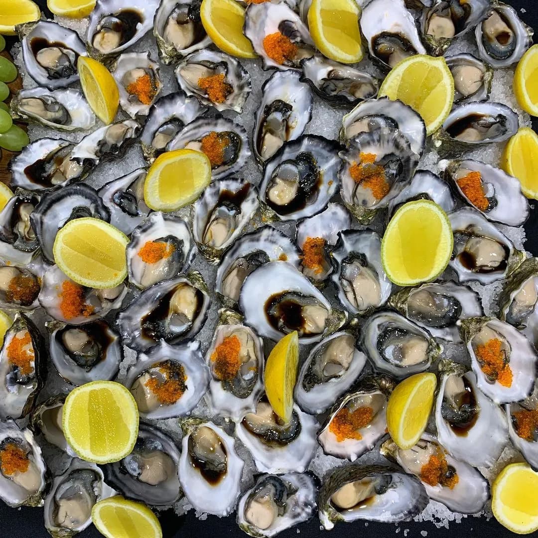 A close-up of numerous oysters on the half shell, topped with bright orange fish roe and lemon slices, arranged on a bed of crushed ice. Green grapes are visible in the top left corner of the image. The oysters appear fresh and ready to eat.