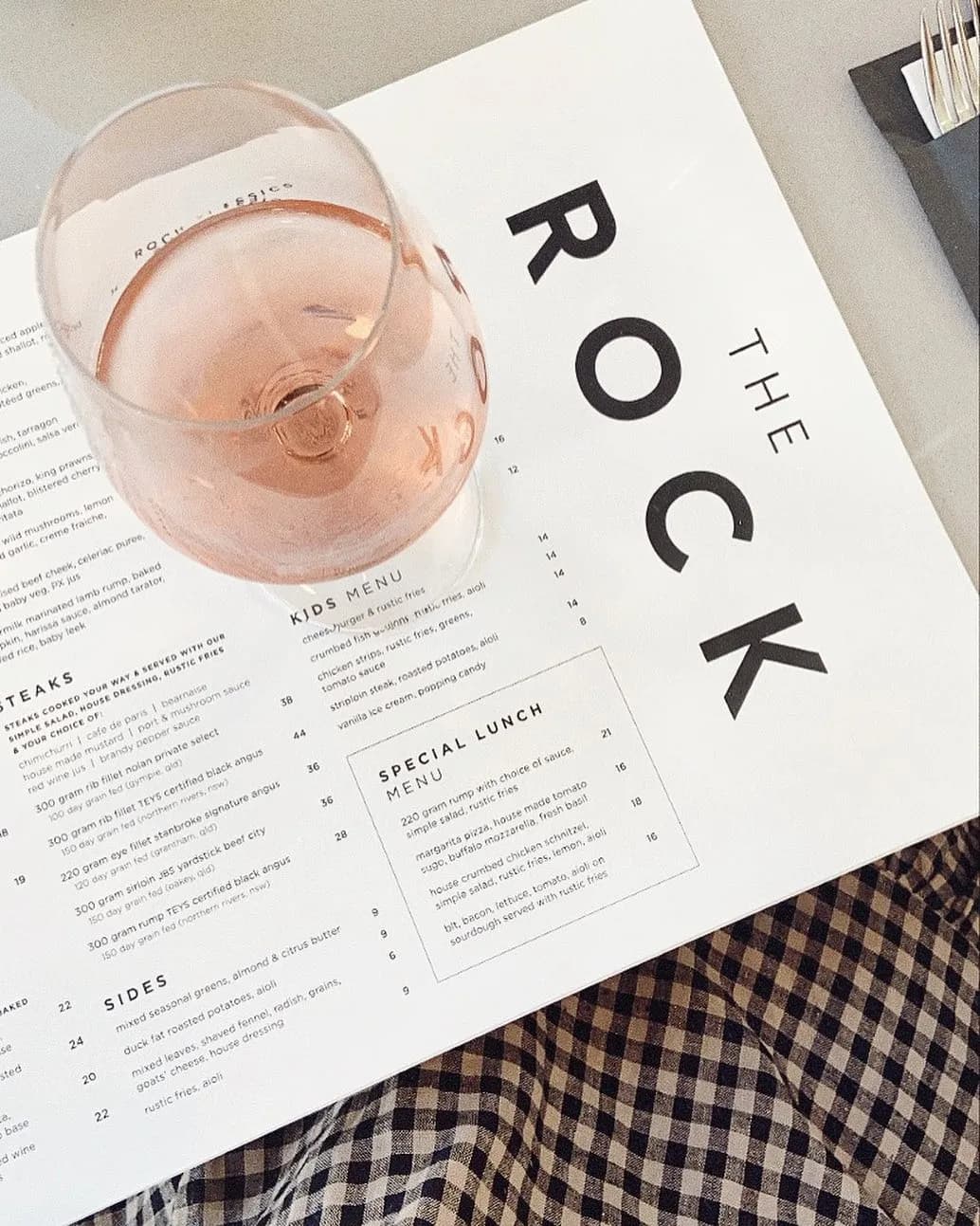 A menu from "The Rock" restaurant rests on a table with a glass of pink rosé wine placed on top. The menu features sections for steaks, sides, kids' menu, and a special lunch menu. Checkered cloth or napkin is partially visible at the bottom of the image.