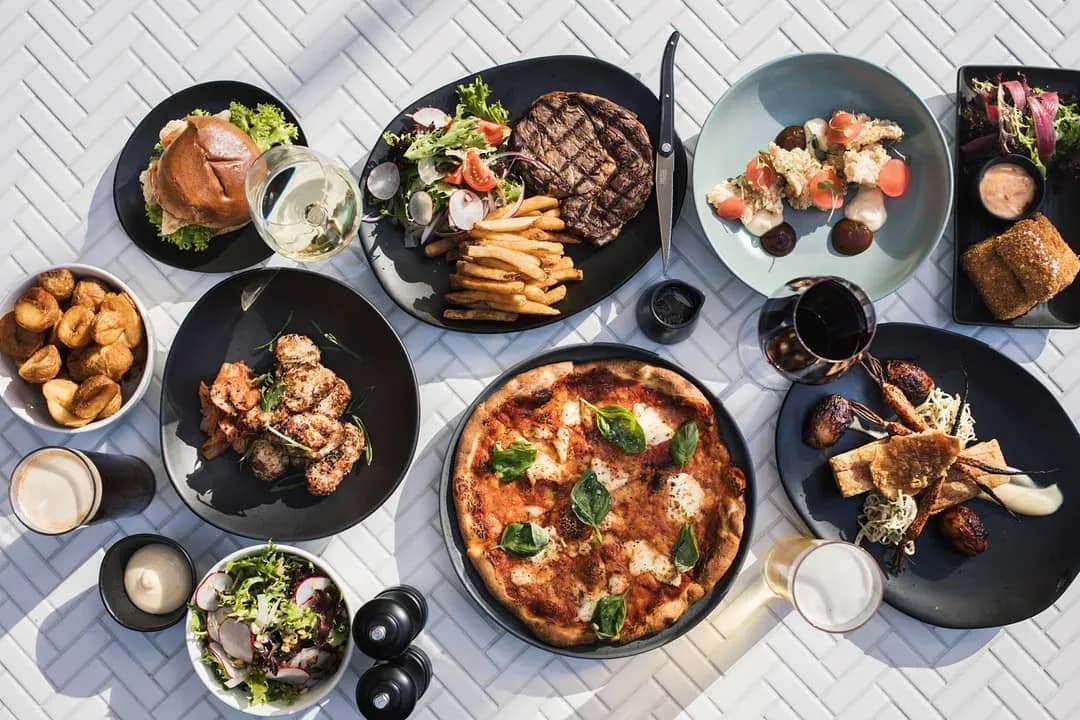 An overhead view of a variety of dishes on a table, including a pizza with basil, a grilled steak with fries, a burger with salad, chicken wings, a bowl of salad, breaded appetizers, a bowl of meatballs, and several drinks, all set against a white textured tablecloth.