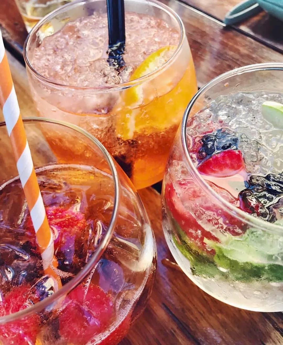 Close-up of three colorful cocktails on a wooden table. The drinks are garnished with various fruits, including orange slices, lime wedges, and berries. Each glass has a straw, with one being striped. The vibrant colors of the drinks stand out against the wood background.