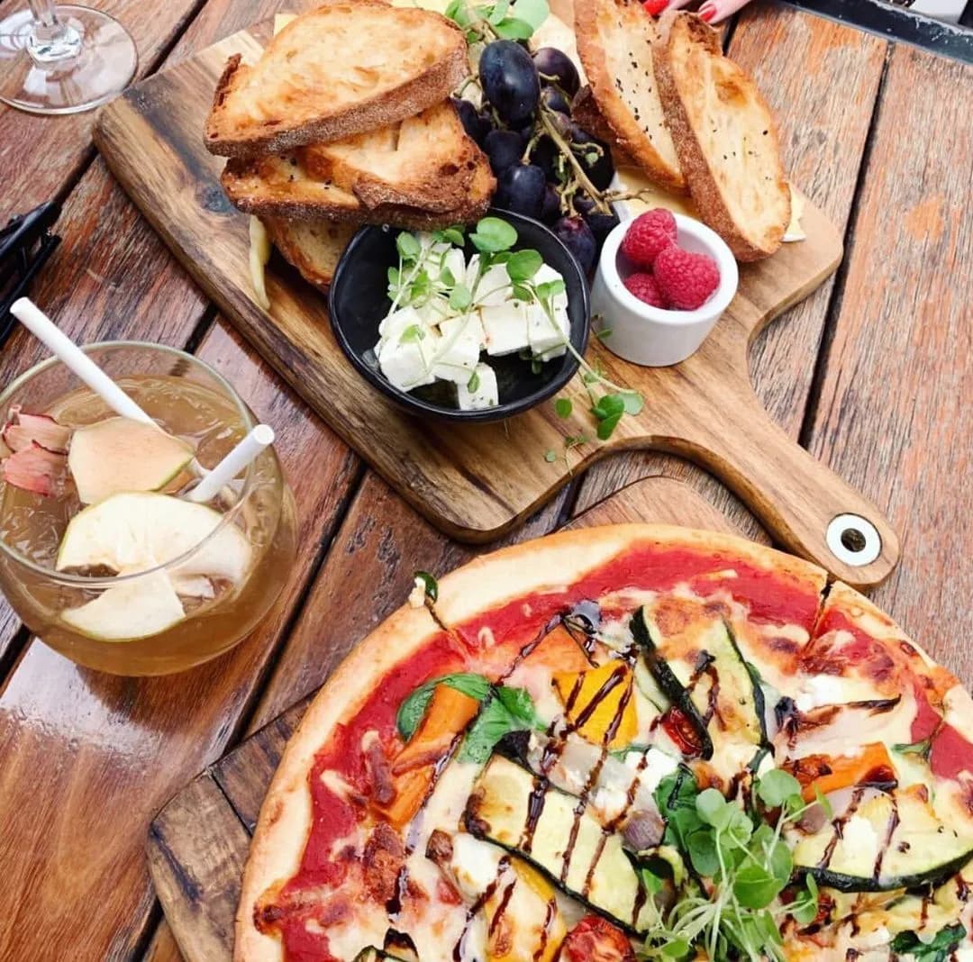 A vibrant spread featuring a vegetable pizza with a variety of colorful toppings, a wooden board with toasted bread slices, a bowl of white cheese cubes, grapes, raspberries, and a cocktail with apple slices and a straw, all on a wooden table.