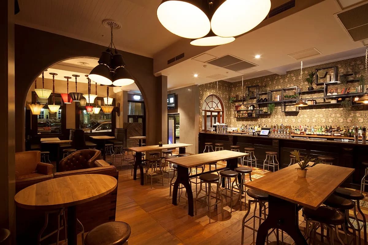 A cozy bar interior featuring a mix of wooden and metal furnishings. Warm, dim lighting and a variety of hanging lamp fixtures create an inviting atmosphere. Shelves stocked with bottles line the bar, and tables with stools are spaced throughout the room.