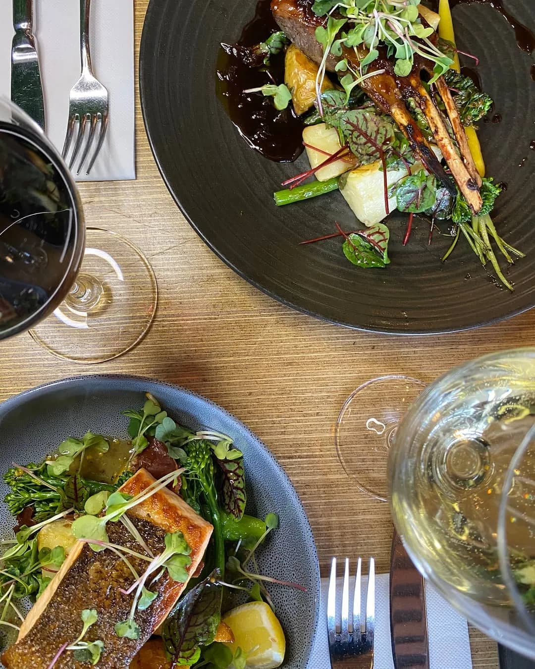 A table setting with two elegant dishes and wine glasses. The dish on the top is garnished with microgreens and roasted vegetables on a dark plate. The dish on the bottom features a piece of fish with microgreens on a blue plate. Red and white wine are served.