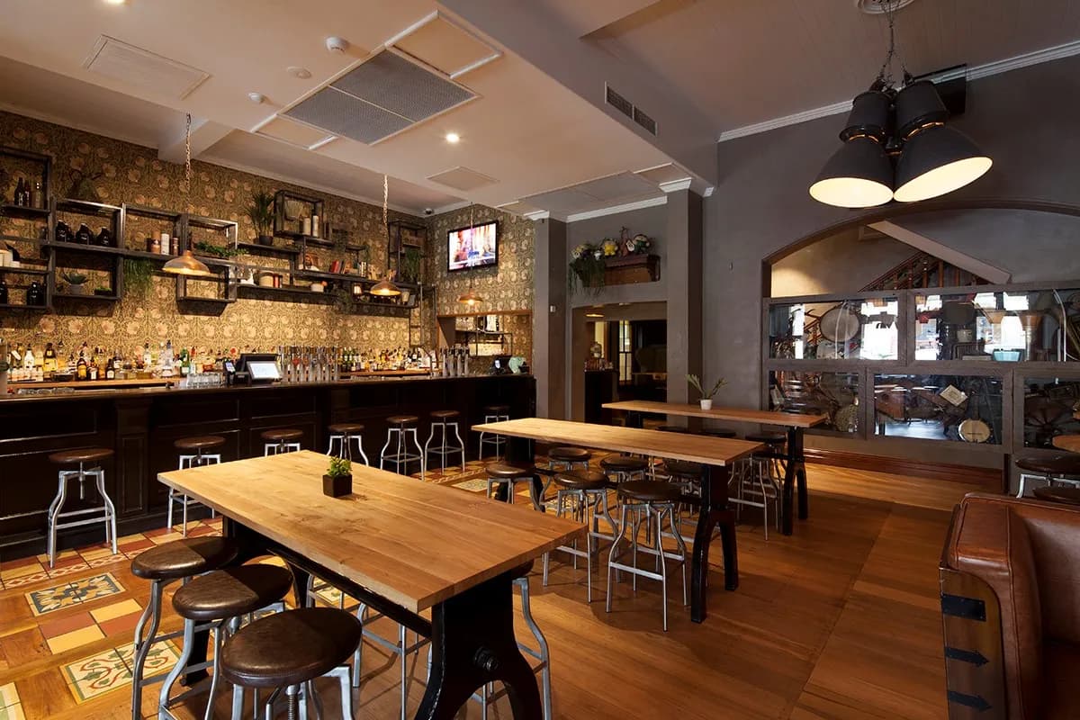 A cozy bar interior with wooden tables and stools, decorative wallpaper, and various bottles displayed on shelves behind the bar. Warm lighting creates a welcoming ambiance. A TV is mounted above the bar displaying a show or advertisement.