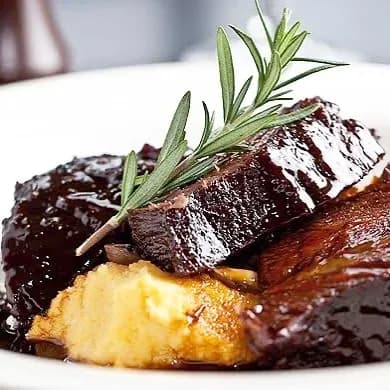 A close-up shot of a gourmet dish featuring tender, dark meat with a glossy sauce glaze, accompanied by a creamy potato puree. The dish is garnished with a sprig of fresh rosemary, enhancing its visual appeal.