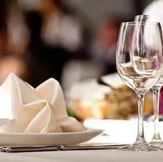 A close-up of a table setting with two empty wine glasses, a folded white napkin on a plate, and blurred background elements suggesting a dining atmosphere.