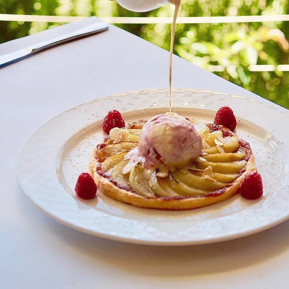 A gourmet dessert on a white plate featuring a fruit tart topped with sliced pears and a scoop of ice cream. Raspberries surround the tart, and a sauce is being elegantly poured over the ice cream. The background shows greenery outside a window.