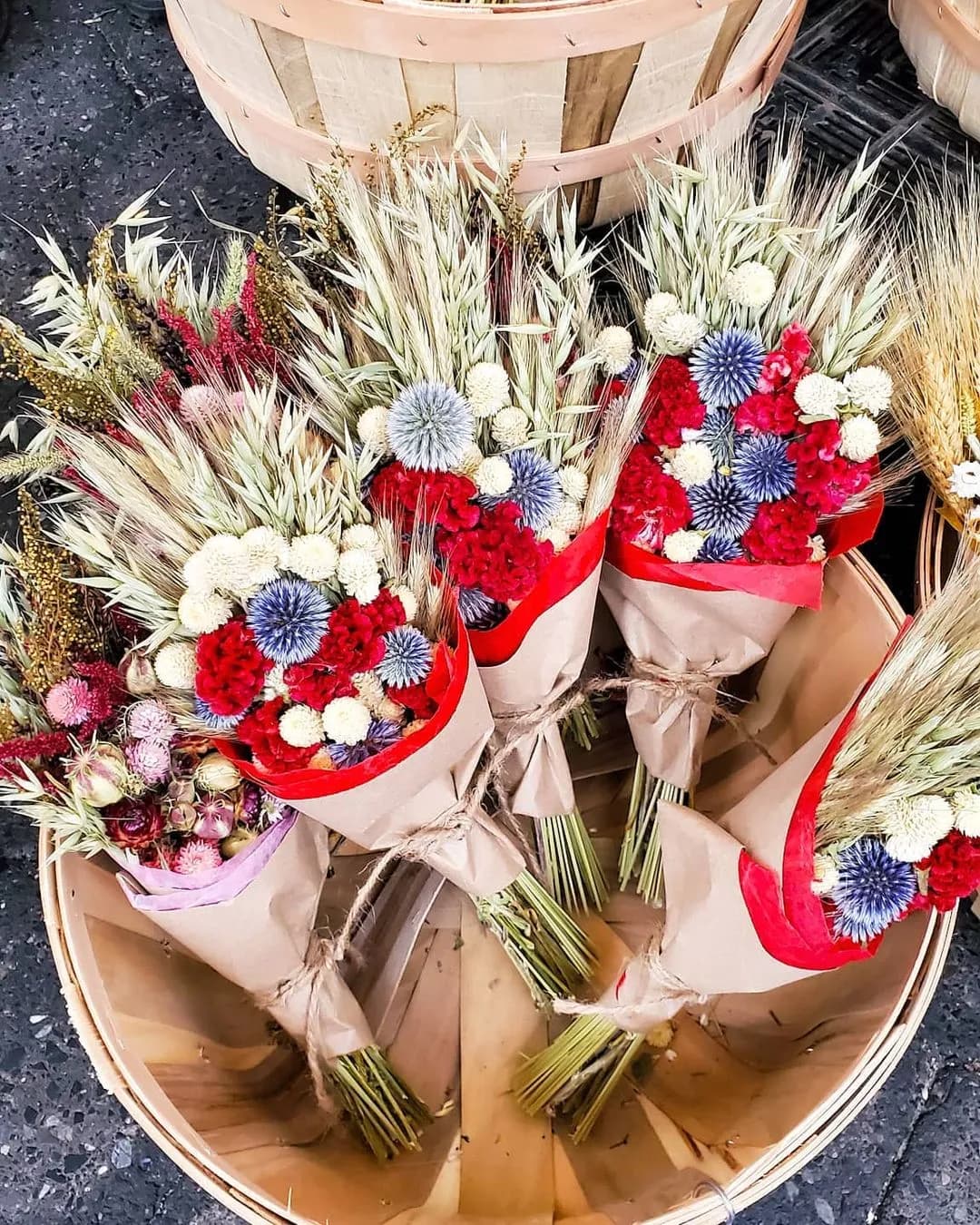 A collection of vibrant dried flower bouquets wrapped in brown paper are displayed in a woven basket. Each bouquet contains a mix of red, white, blue, and green flowers, accentuated with tall, dried grasses for a rustic look.