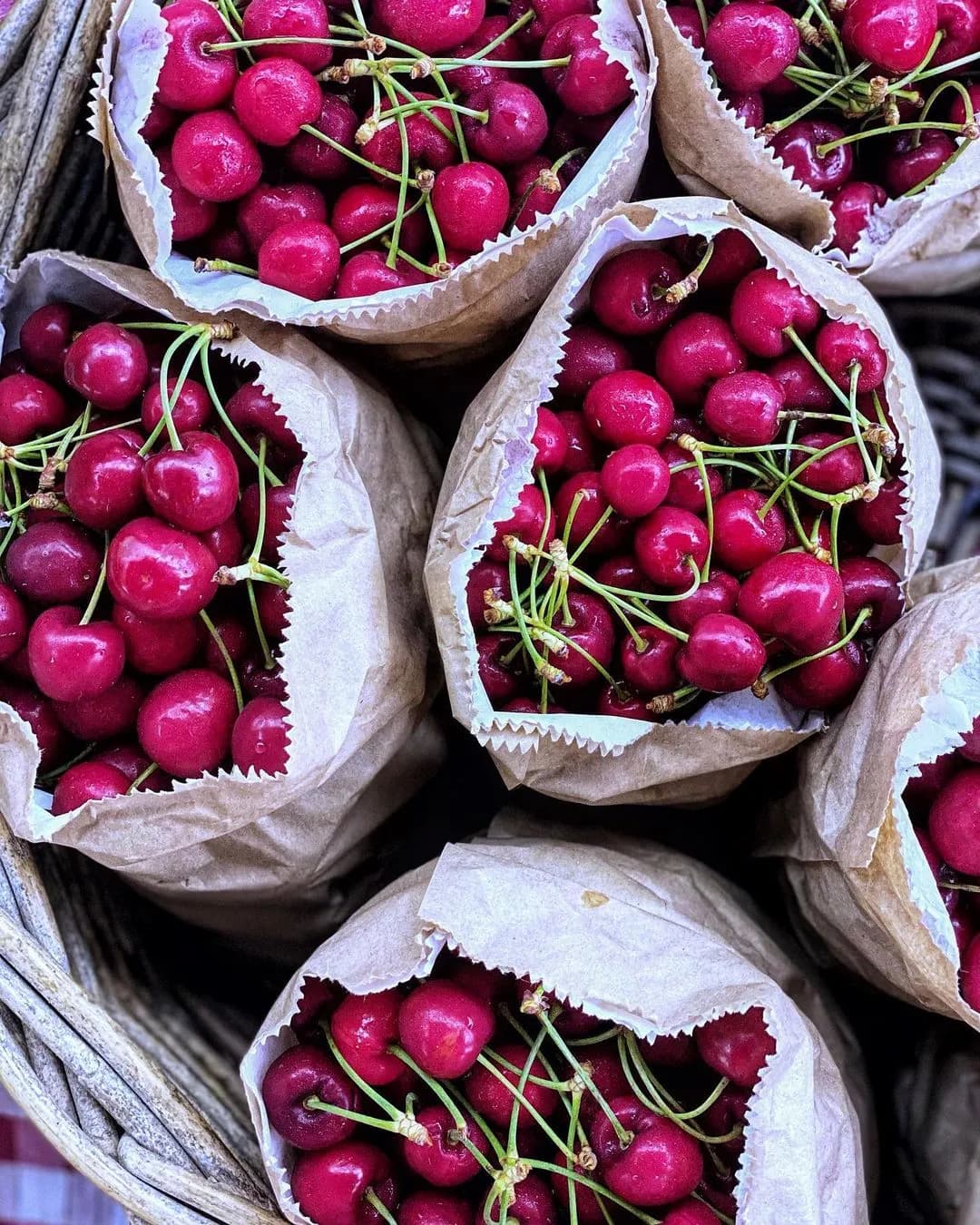 Bags of vibrant red cherries with green stems are arranged in a basket. The cherries fill the paper bags to the brim, showcasing their freshness and juiciness.