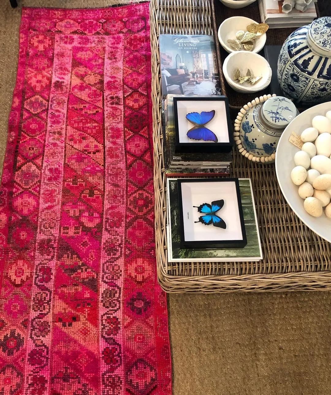 A vibrant pink patterned rug lies on a carpeted floor. Adjacent is a wicker table displaying various items, including books, blue butterfly art frames, bowls, and decorative blue-and-white ceramics, including a tea set and a bowl of white spheres.