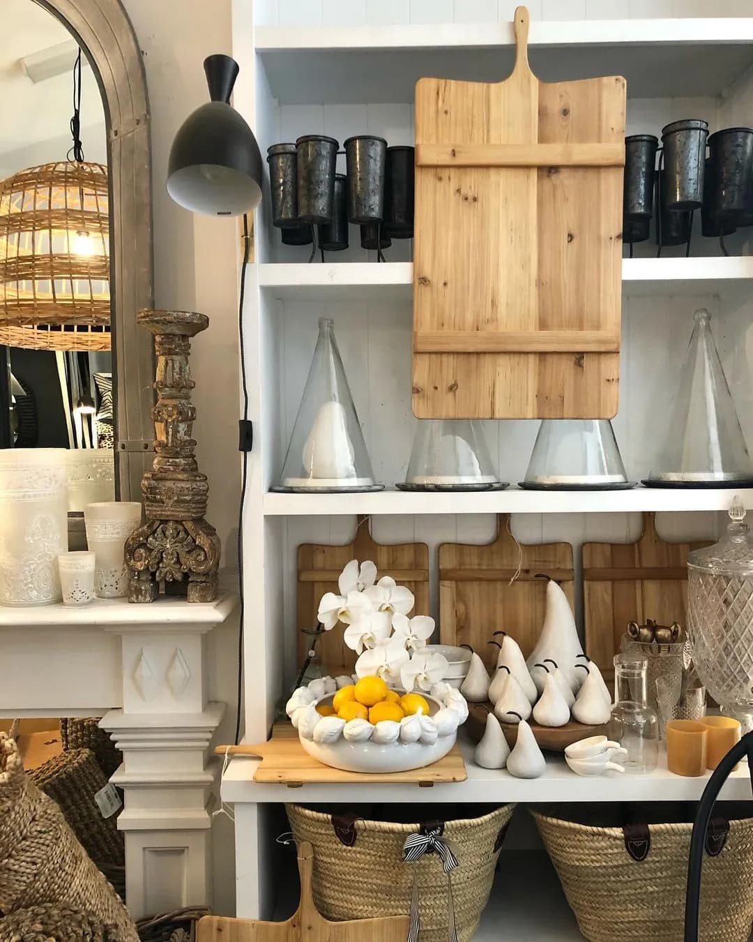 A cozy interior showcases a rustic shelf with wooden cutting boards, glass flasks, black candles, and white ceramic pears. A bowl of lemons and white orchids sits on the bottom shelf. Natural woven baskets and vintage decor items are arranged nearby.