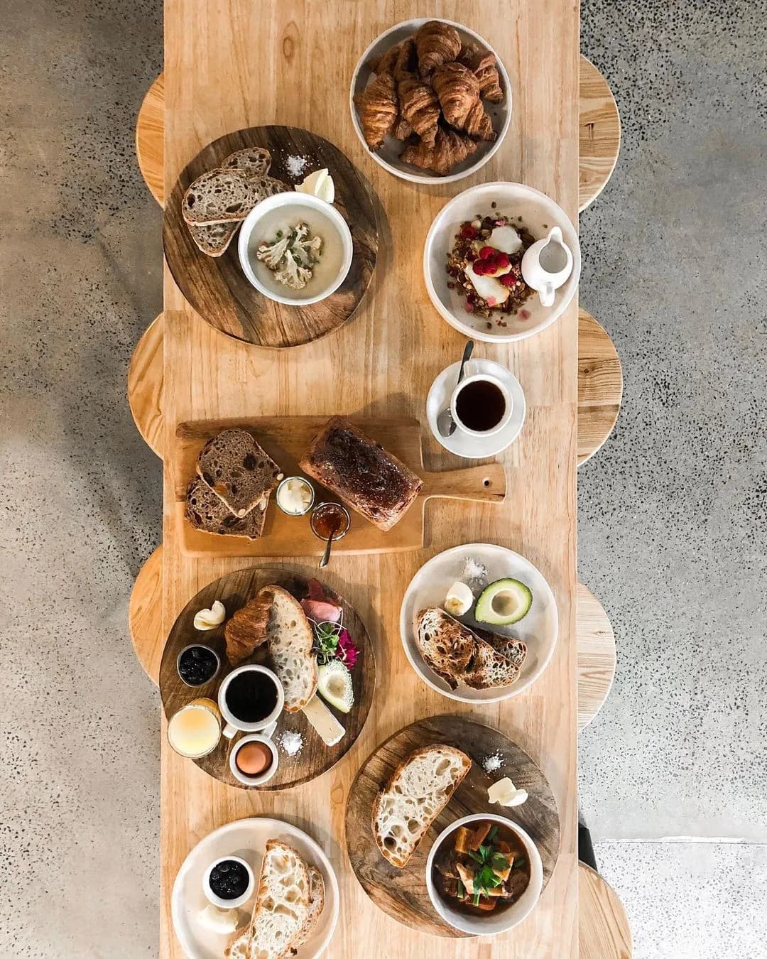 A wooden table set with various breakfast items, including croissants, yogurt with fruit, coffee, assorted bread slices with butter, avocado, black jam, soft-boiled eggs, and bowls with granola and fresh berries. The table is viewed from above.