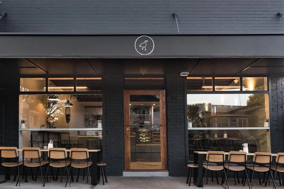 A modern café exterior with a dark facade, large windows, and a wooden door in the center. The café has outdoor seating with wooden chairs and tables arranged neatly. The café sign above has a simple bird logo. The reflections in the windows show nearby buildings.