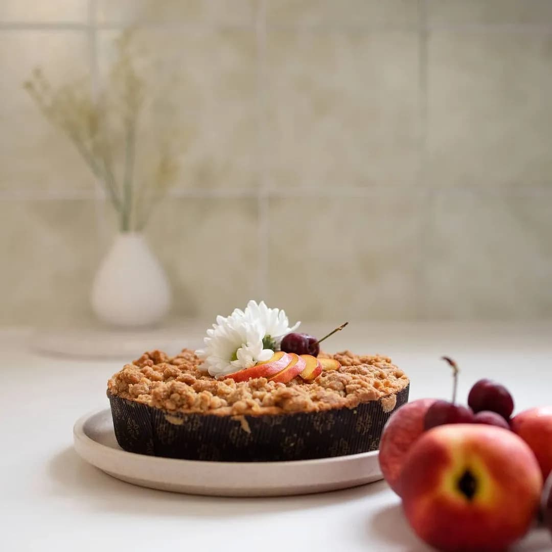 A crumbly pie adorned with a white flower, peach slices, and cherries sits on a plate in a minimalist kitchen. In the foreground, fresh peaches and cherries are displayed. The background features a simple vase with delicate dried flowers on a tiled countertop.