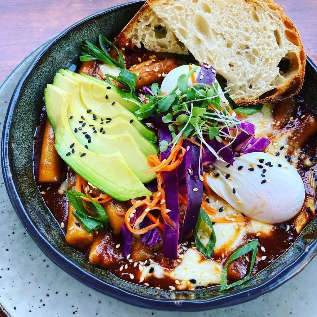 A bowl of spicy Korean rice cakes (tteokbokki) topped with avocado slices, poached eggs, shredded purple cabbage, carrot strips, fresh herbs, and black sesame seeds. A piece of crusty bread is placed on the edge of the bowl.