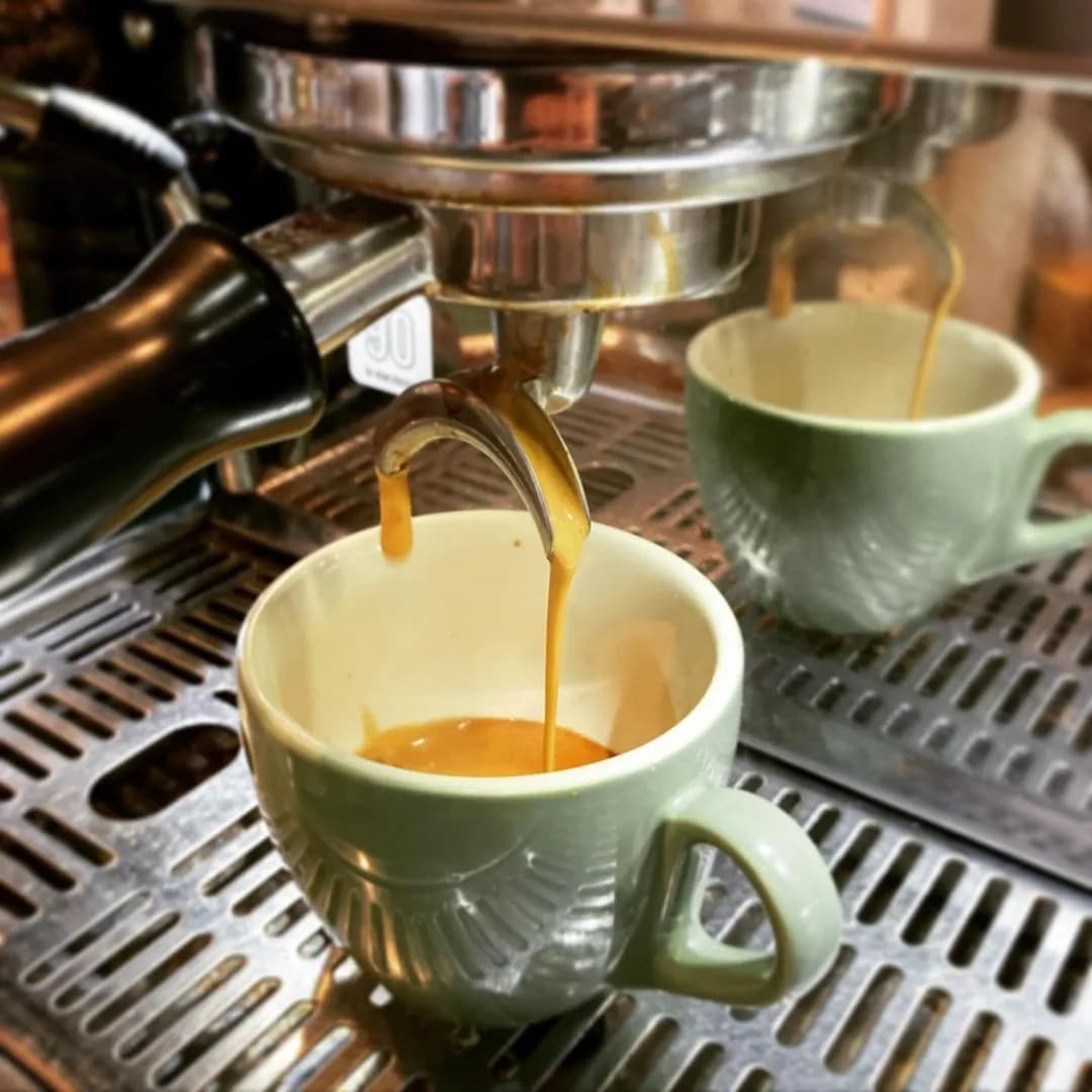 A close-up shot of an espresso machine brewing coffee into a light green ceramic cup. The espresso is streaming out of the portafilter, creating a rich golden crema in the cup. The setting appears to be a kitchen or cafe.