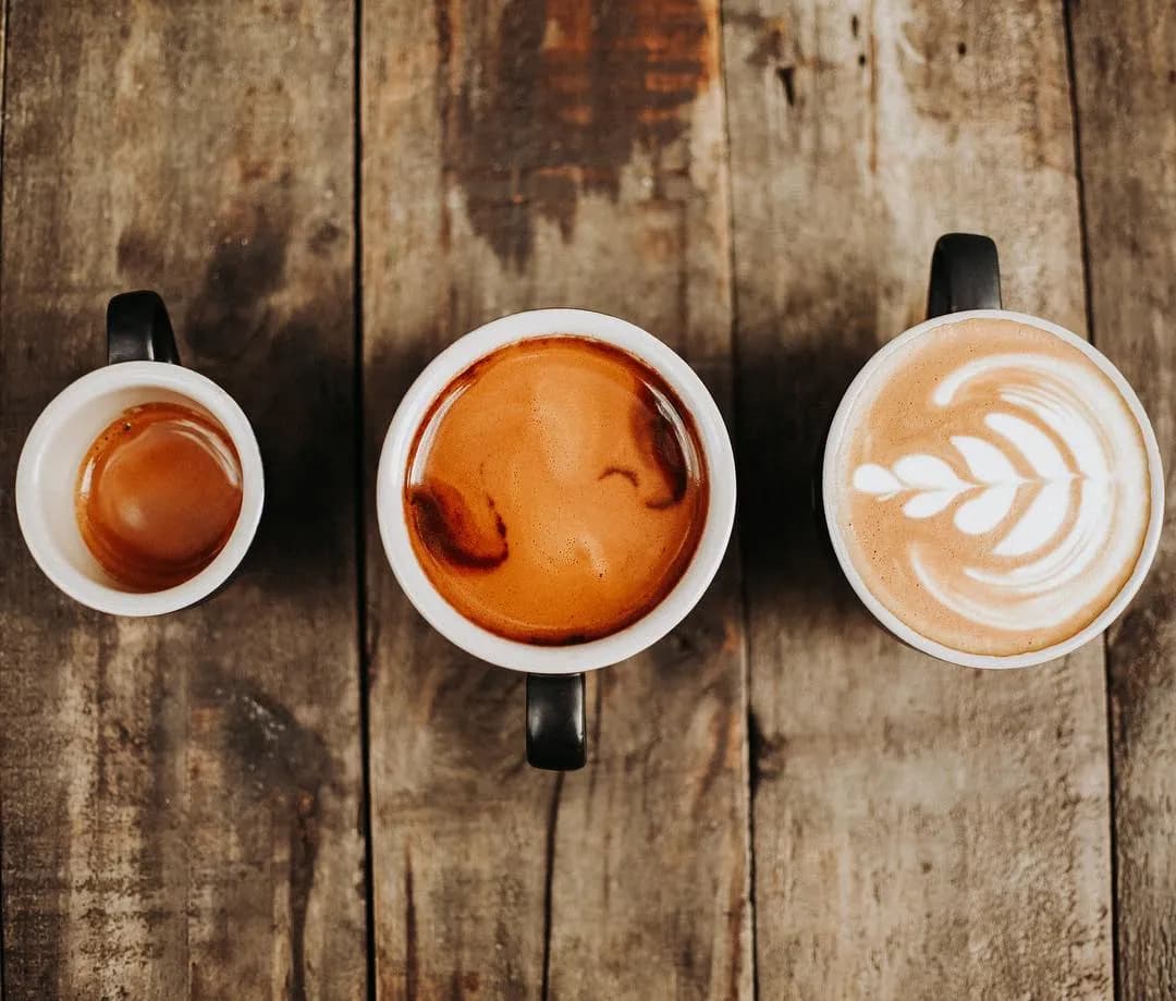 A top-down view of three coffee cups placed on a rustic wooden surface. From left to right, there is a small cup of espresso, a medium cup of espresso with crema, and a large cup of latte with latte art in the shape of a leaf.