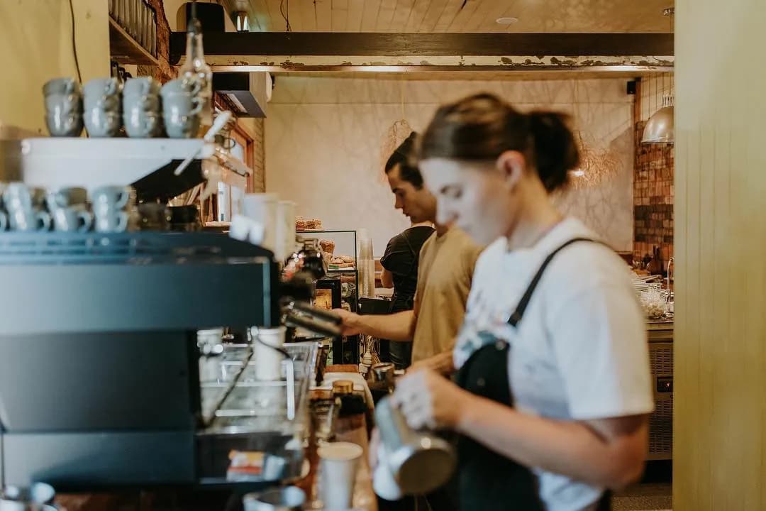 Two baristas work behind a counter in a cozy café. The woman in the foreground froths milk, while the man in the background operates an espresso machine. Shelves with cups and a warm, rustic interior decor are visible. The scene exudes a busy but welcoming atmosphere.