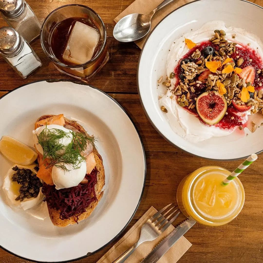 A wooden table is set with a breakfast spread. There is toast with poached eggs, shredded beets, and greens on one plate, a bowl of yogurt topped with fruit and granola, a glass of iced coffee, and a glass of orange juice with a green-striped straw.