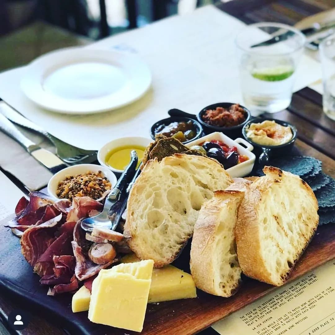 A wooden platter with assorted cheeses, slices of bread, cold cuts, and various condiments in small bowls is set on a table. A glass of water with lime, a white plate, cutlery, and napkins are also visible on the table.