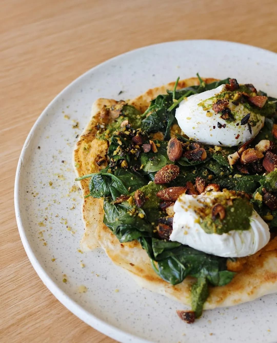 A plate of food featuring leafy greens, two poached eggs, and a sprinkling of nuts on a piece of flatbread, all garnished with a green sauce and spices. The plate rests on a light wooden table.