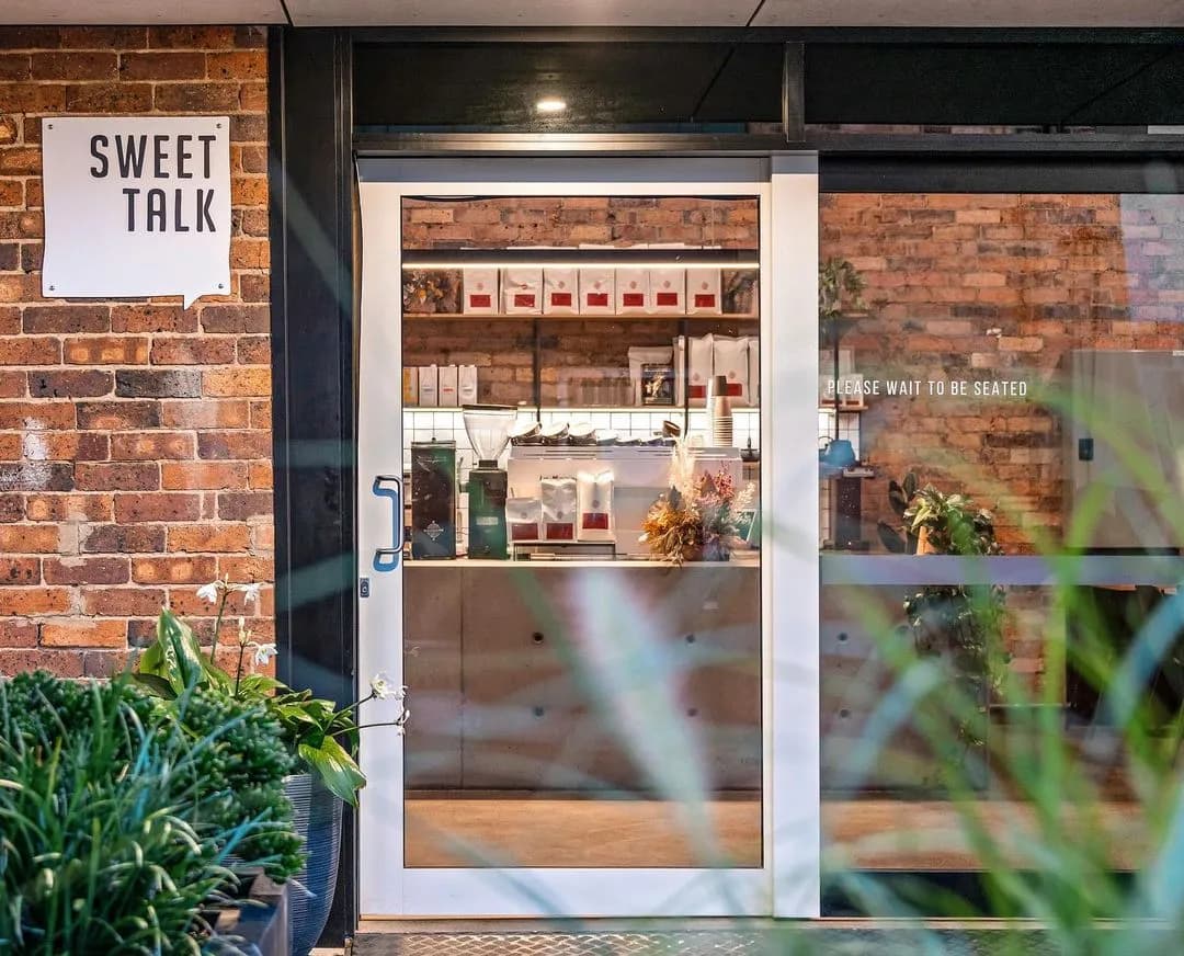 Entrance of a café named "Sweet Talk" with a sign on a brick wall on the left. The glass door shows the interior with a counter and shelves holding various items. A sign on the door reads, "Please wait to be seated." Plants frame the entrance.