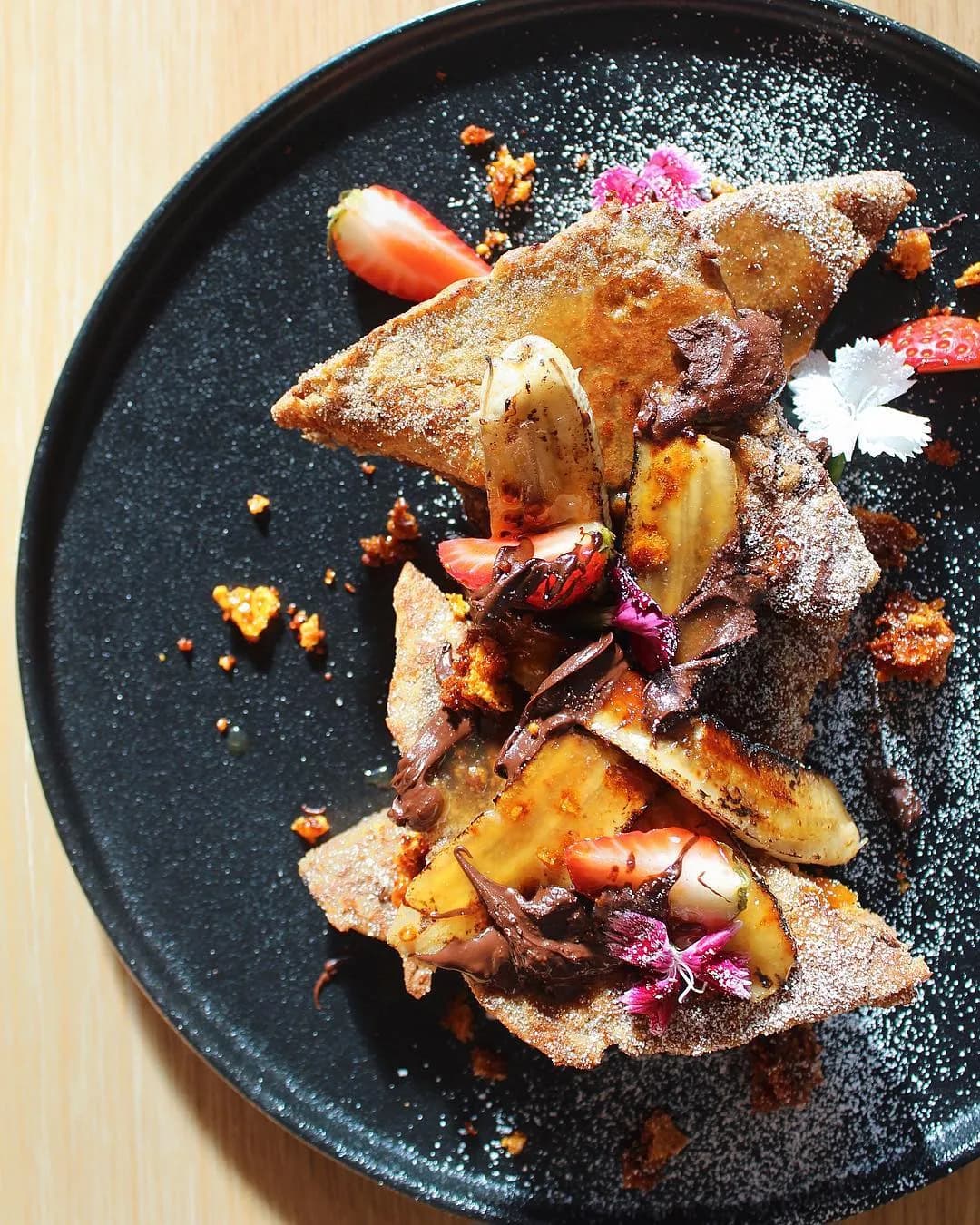 A plate of French toast topped with banana slices, strawberries, chocolate spread, and edible flowers. The dish is dusted with powdered sugar and garnished with crunchy caramel pieces. It is served on a black plate against a light wooden background.