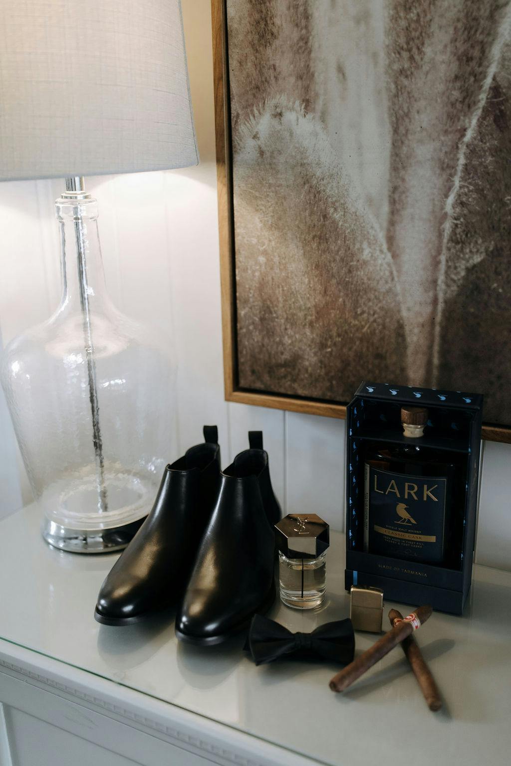 A neatly arranged dresser features black dress boots, a bow tie, gold cufflinks, cigars, a bottle of cologne, a boxed bottle of Lark whiskey, and a clear glass lamp with a white shade. A framed abstract art piece hangs above the dresser.