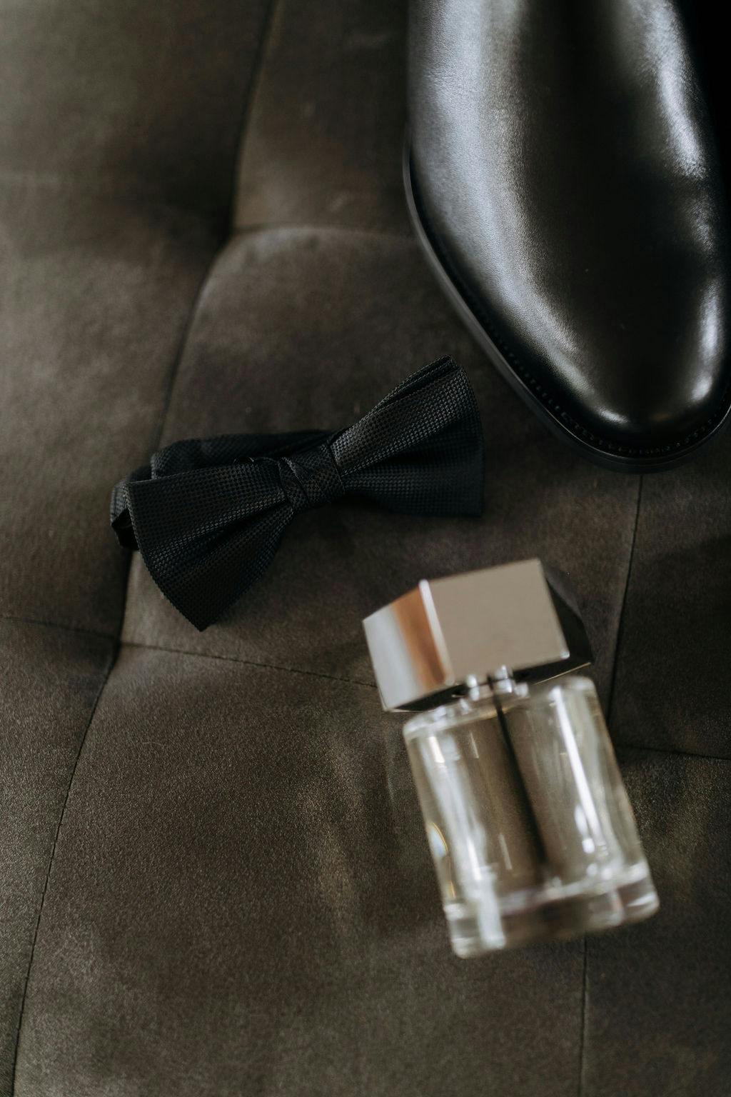 Close-up of a black bow tie, a clear square glass bottle with a silver cap, and the toe of a polished black dress shoe placed on a dark, tufted fabric surface.