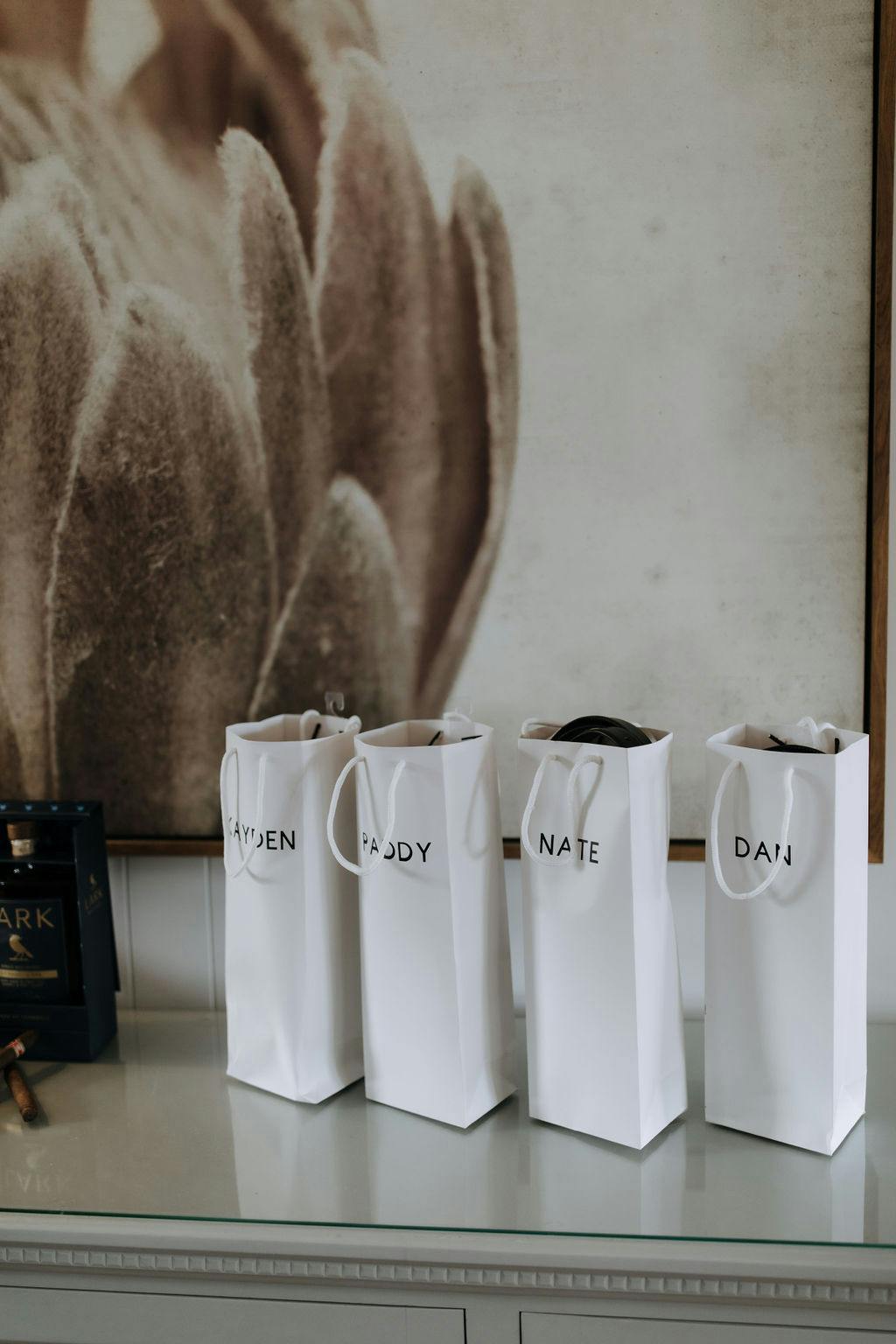 Four white gift bags with names (Jayden, Paddy, Nate, Dan) written on them are lined up on a glass table. A large, framed artwork with soft, neutral colors hangs on the wall behind the bags. A black box is partially visible on the left side of the table.