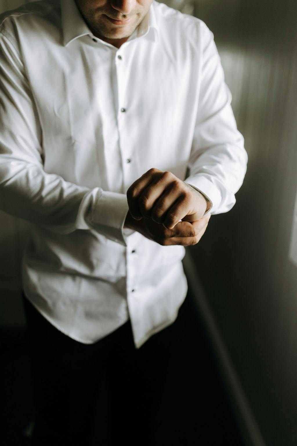 A man in a white dress shirt is adjusting his cufflinks. The focus is on his hands and shirt, with the upper part of his face not visible. He is standing in a room with soft, natural light coming from the right side.