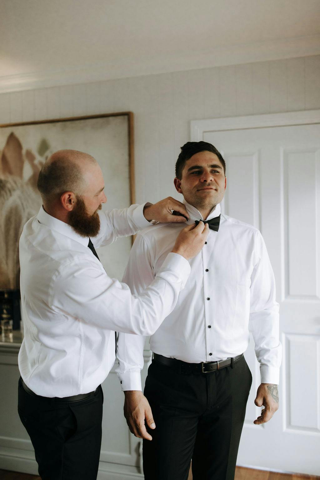 A bearded man helps another man adjust his black bow tie. Both are dressed in white shirts and black pants. They stand in a well-lit room with a white door and a painting in the background. The man being helped looks pleased as he prepares to get dressed.