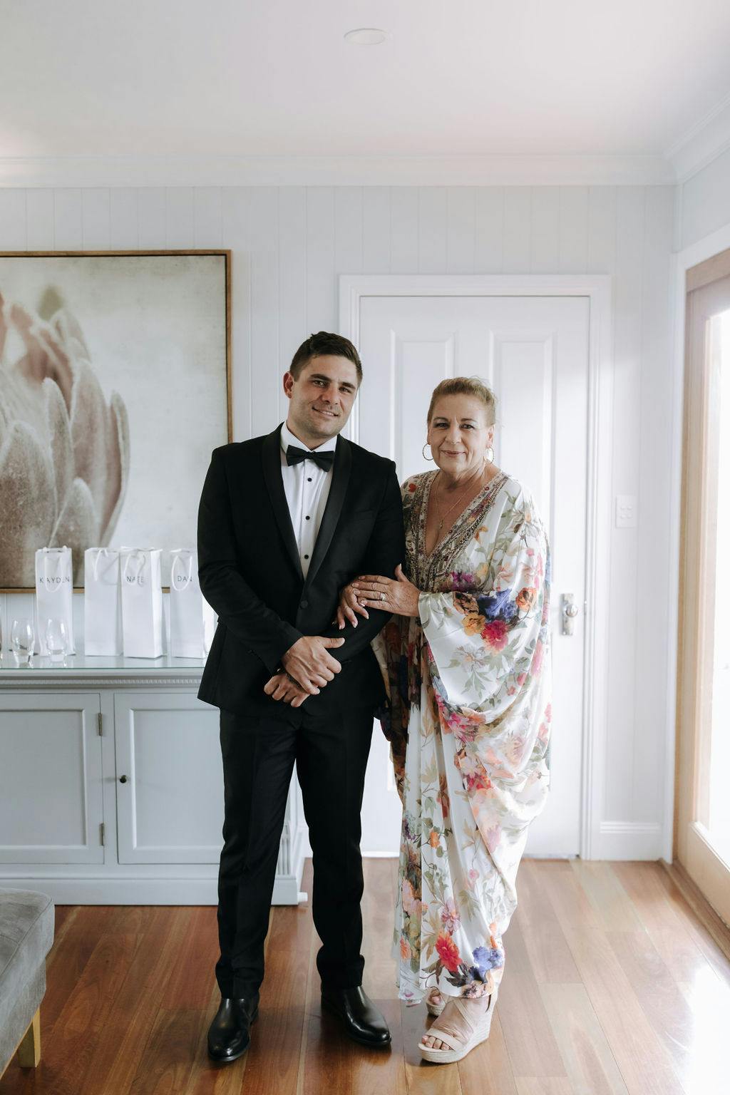 A man in a black tuxedo stands arm-in-arm with a woman in a floral dress. They are indoors in a well-lit room with wooden floors and white walls, with a cabinet and some gift bags behind them. Both are smiling towards the camera.