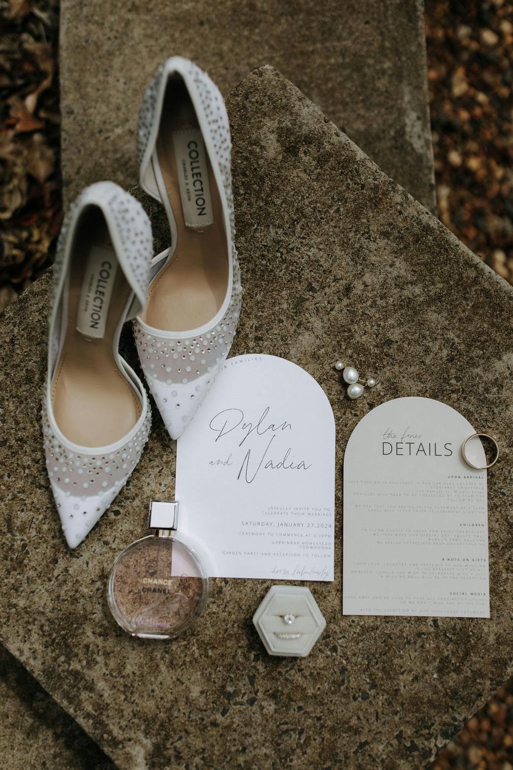 A pair of white, embellished high-heeled shoes is placed on a stone surface next to a wedding invitation that reads "Dylan and Nadia," pearl earrings, a ring, a small perfume bottle, a hexagonal ring box, and a wedding event details card.