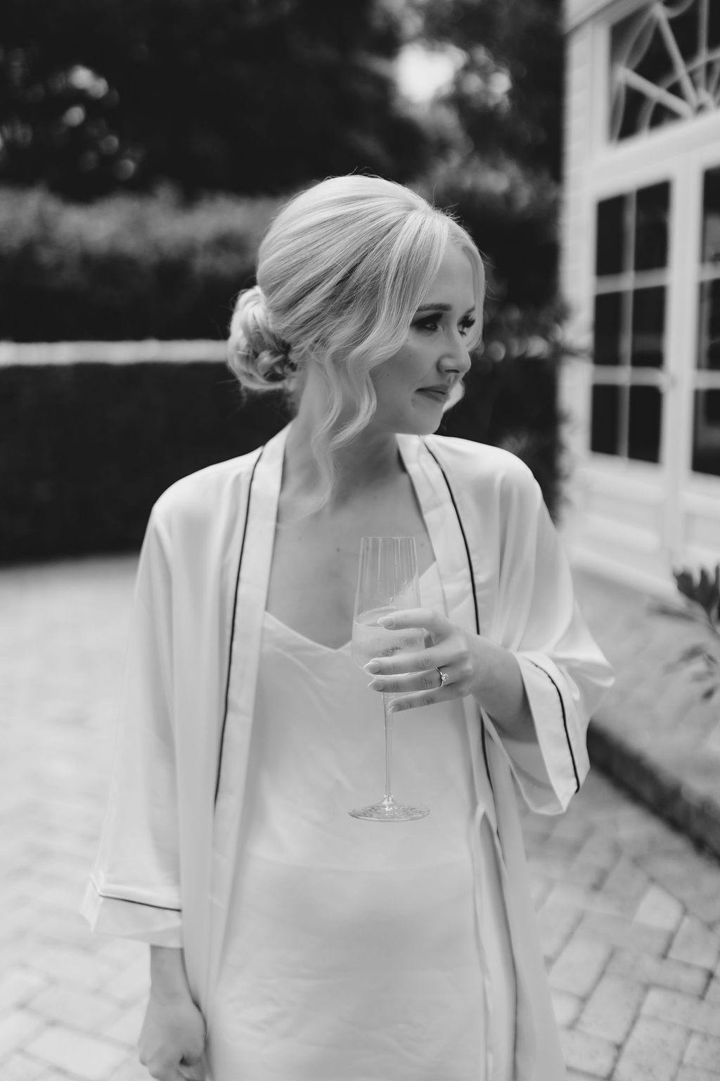 Black and white photo of a person with light hair tied back, wearing a robe and holding a glass of what appears to be champagne. The background features a pathway leading to a house with large windows and some greenery. The person is looking off to the side.