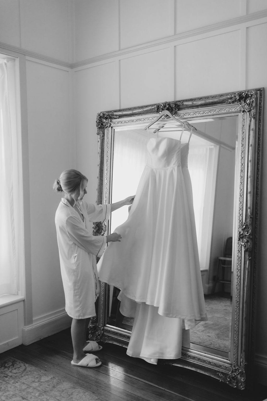 A woman dressed in a robe and slippers stands in a room, adjusting a long, white wedding dress hanging on a hanger in front of a large, ornate mirror. The room has wooden floors and is softly lit by natural light coming through a window. (Black and white image).