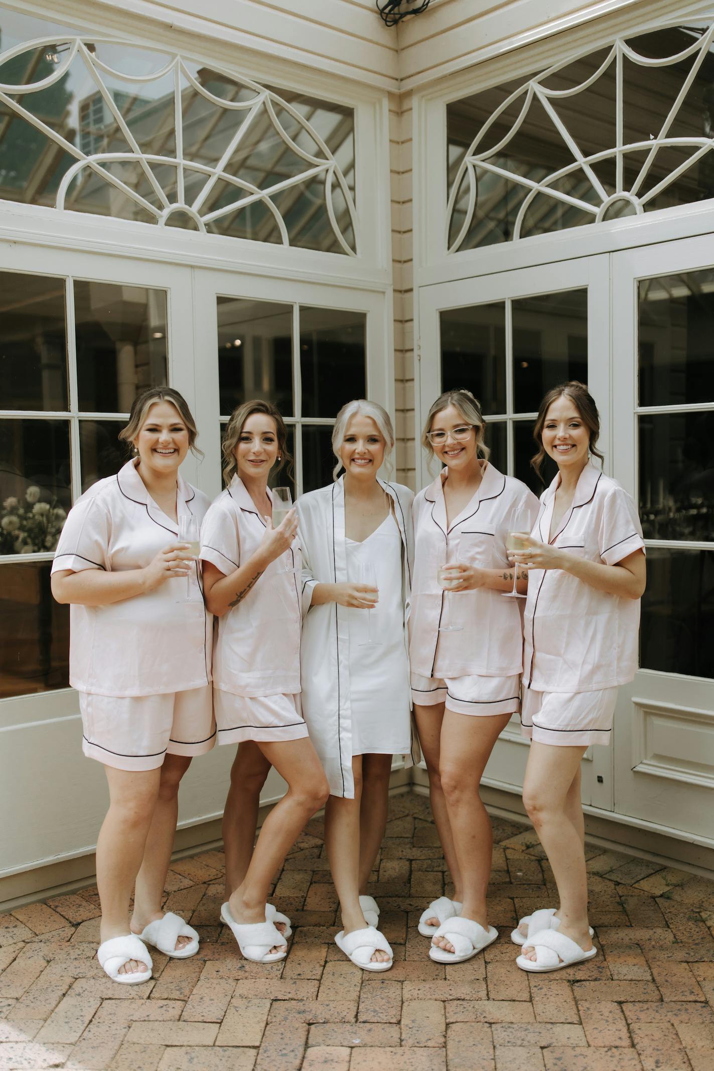 A group of five women wearing matching light pink pajamas and white slippers stand together, smiling and holding champagne glasses. One woman in the center is dressed in a white robe and slippers. Behind them is a building with large windows.